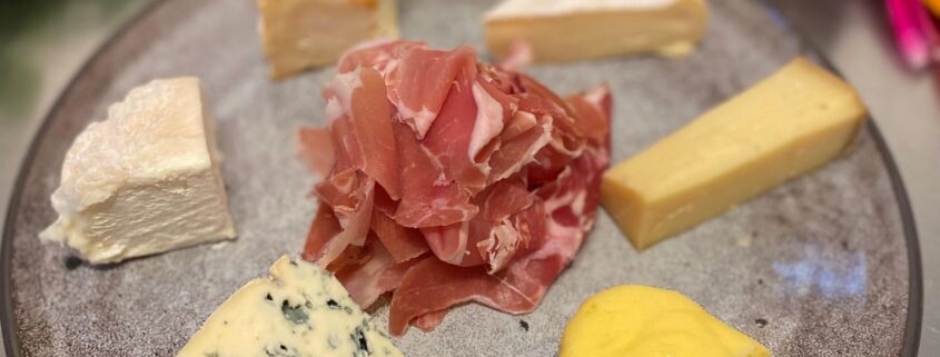 Teller mit sechs verschiedenen Käsesorten, darunter Blauschimmelkäse, und in der Mitte aufgeschichtetem rohem Schinken